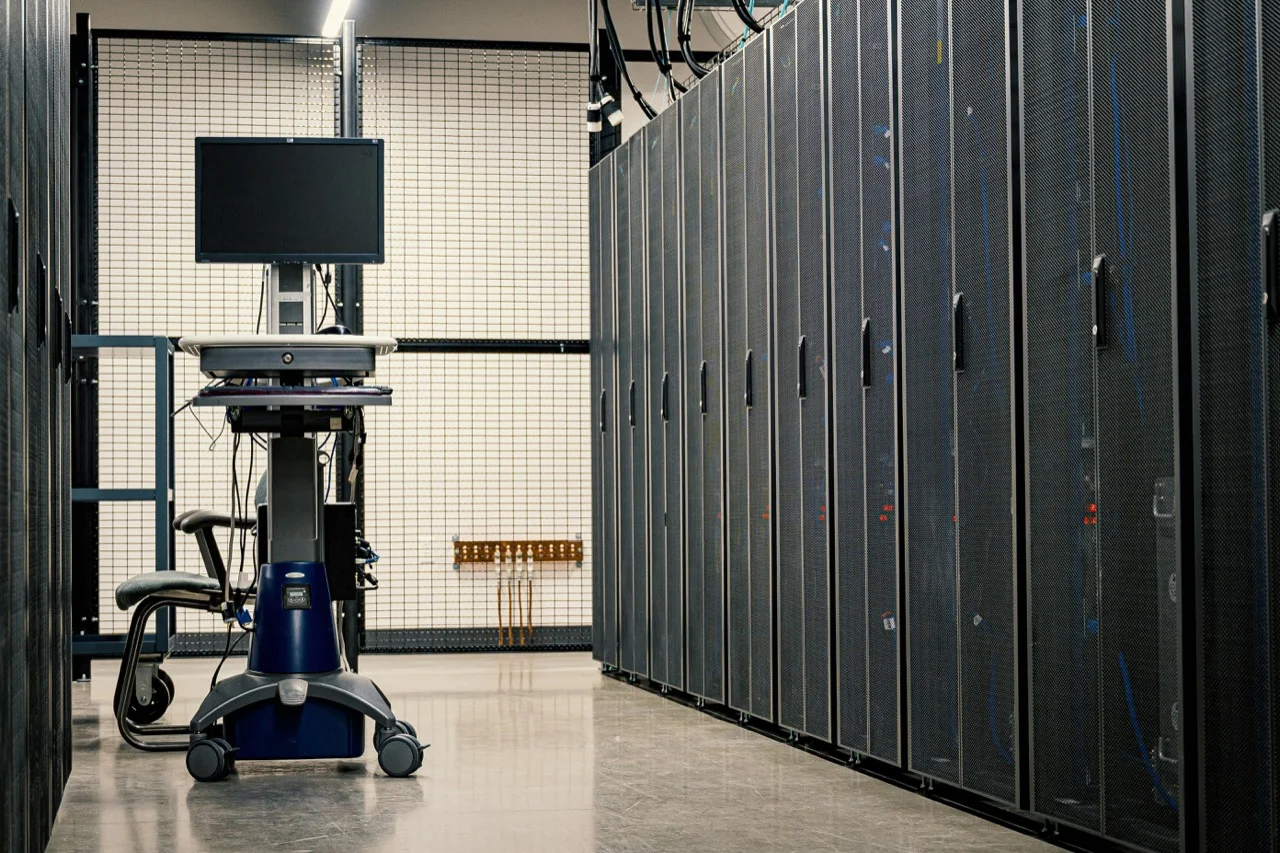 Data-center corridor with server racks and a mobile terminal, representing compute scarcity and technical adaptation under constraint.